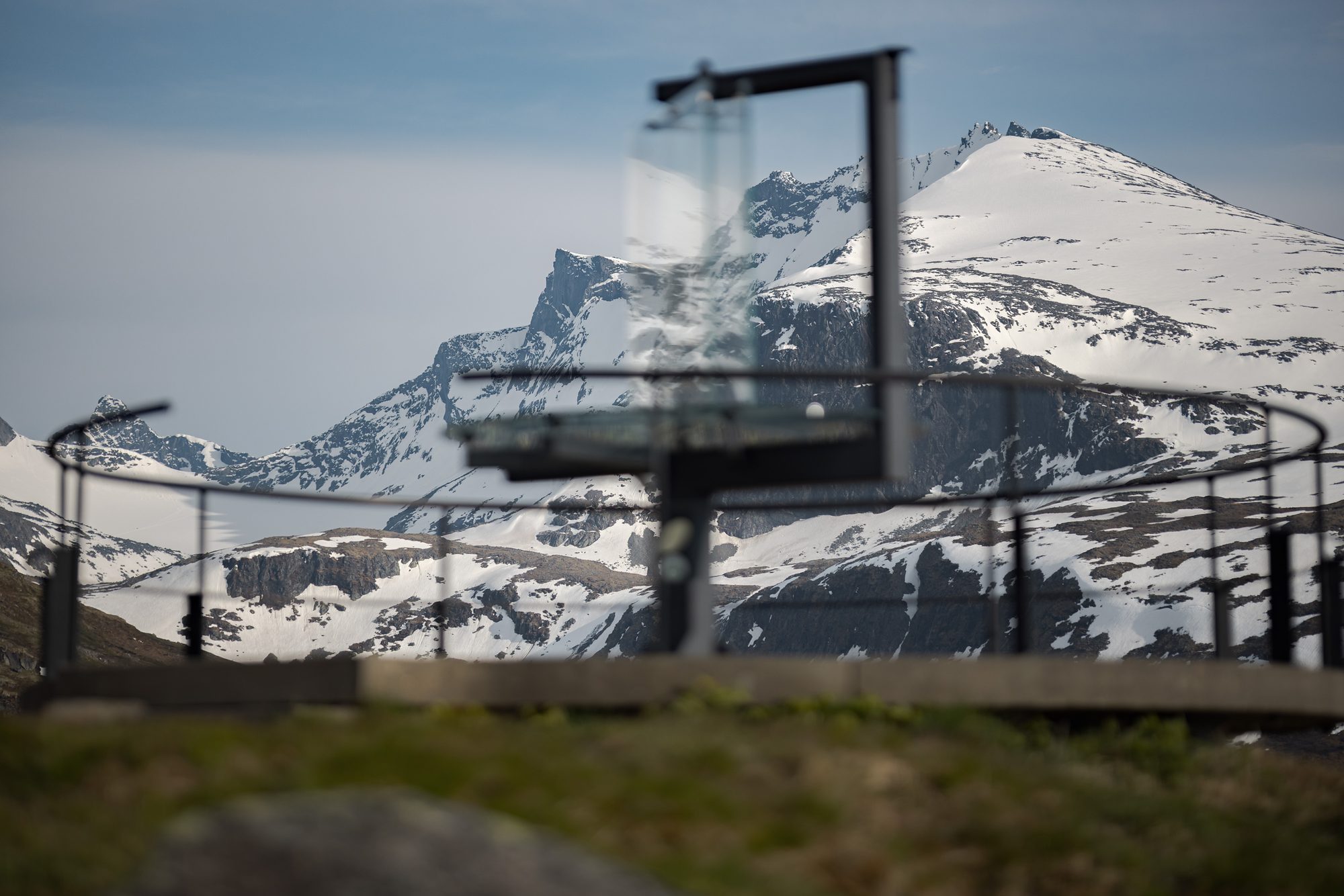 The glass panorama marker at the Oscarshaug viewpoint, its railing and platform in front of the snow-covered peaks of the Hurrungane