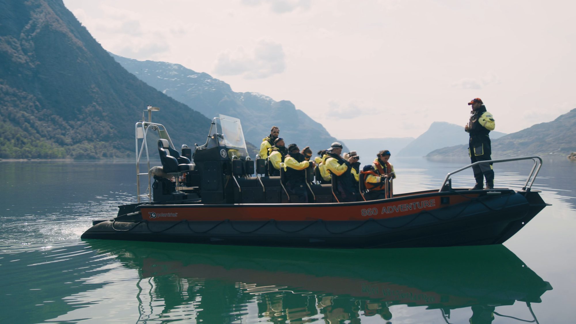 A RIB with a dozen guests in yellow flotation suits resting on still water, the guide standing up in the boat, the fjord and its mountains stretching away behind