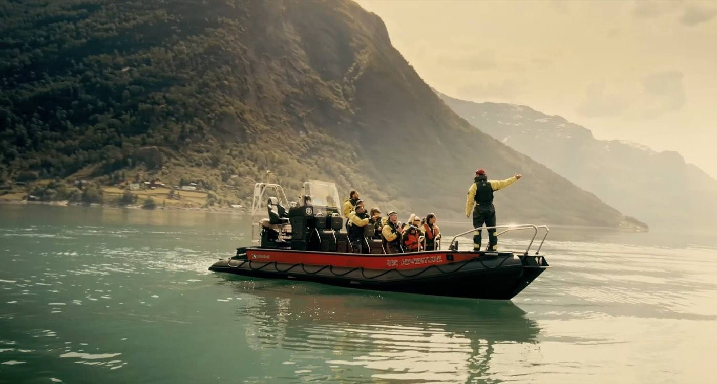 A RIB carrying about a dozen guests in flotation suits across a calm fjord, the guide at the stern pointing out at the water
