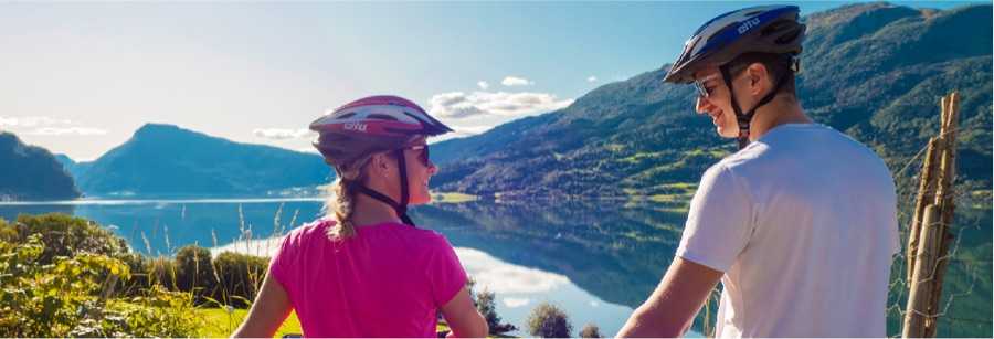 A woman in pink and a man in white, both wearing cycling helmets, seen from behind as they look out over the mirror-still fjord from a garden slope