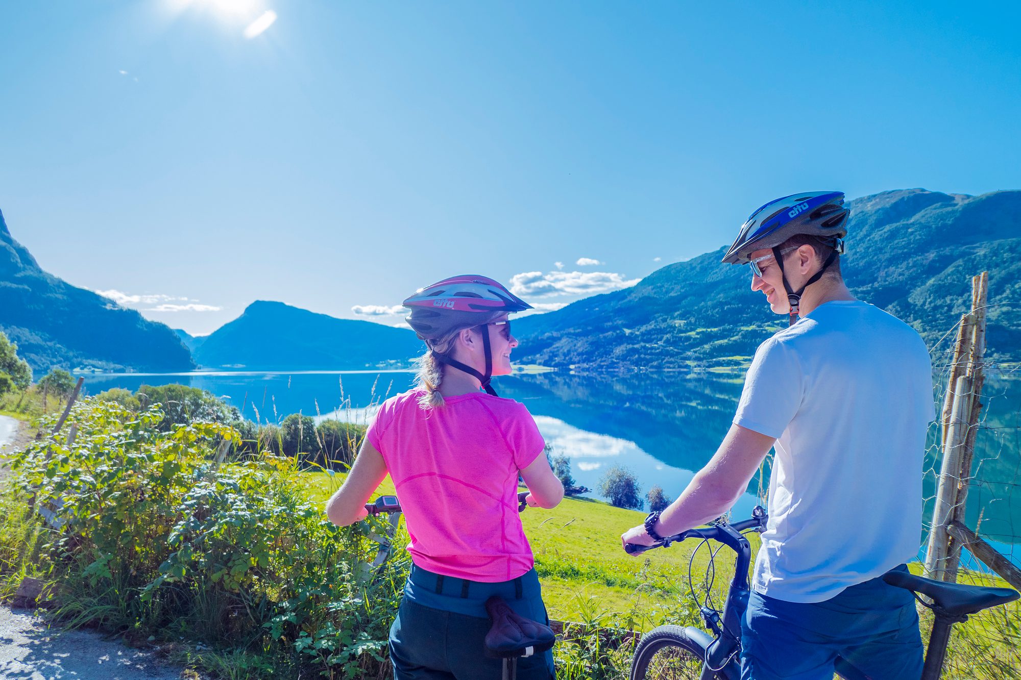 Two cyclists in helmets stopped at the roadside, seen from behind, looking out over the still fjord and the mountains reflected in it