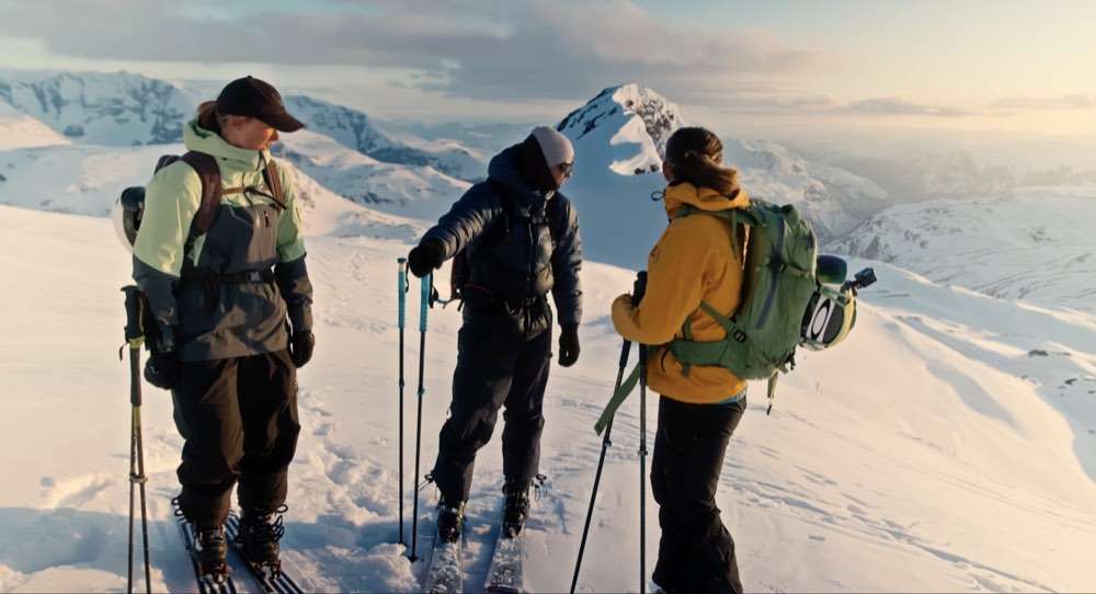 Three ski tourers with skins and packs standing on a snow ridge at sunrise, endless white peaks behind them