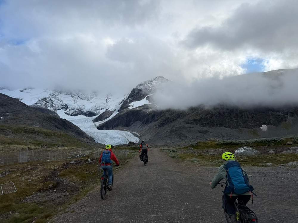 Three riders on electric bikes going away up a gravel track towards the glacier, its tongue spilling white between grey moraine slopes, with cloud lying halfway down the mountainside above them