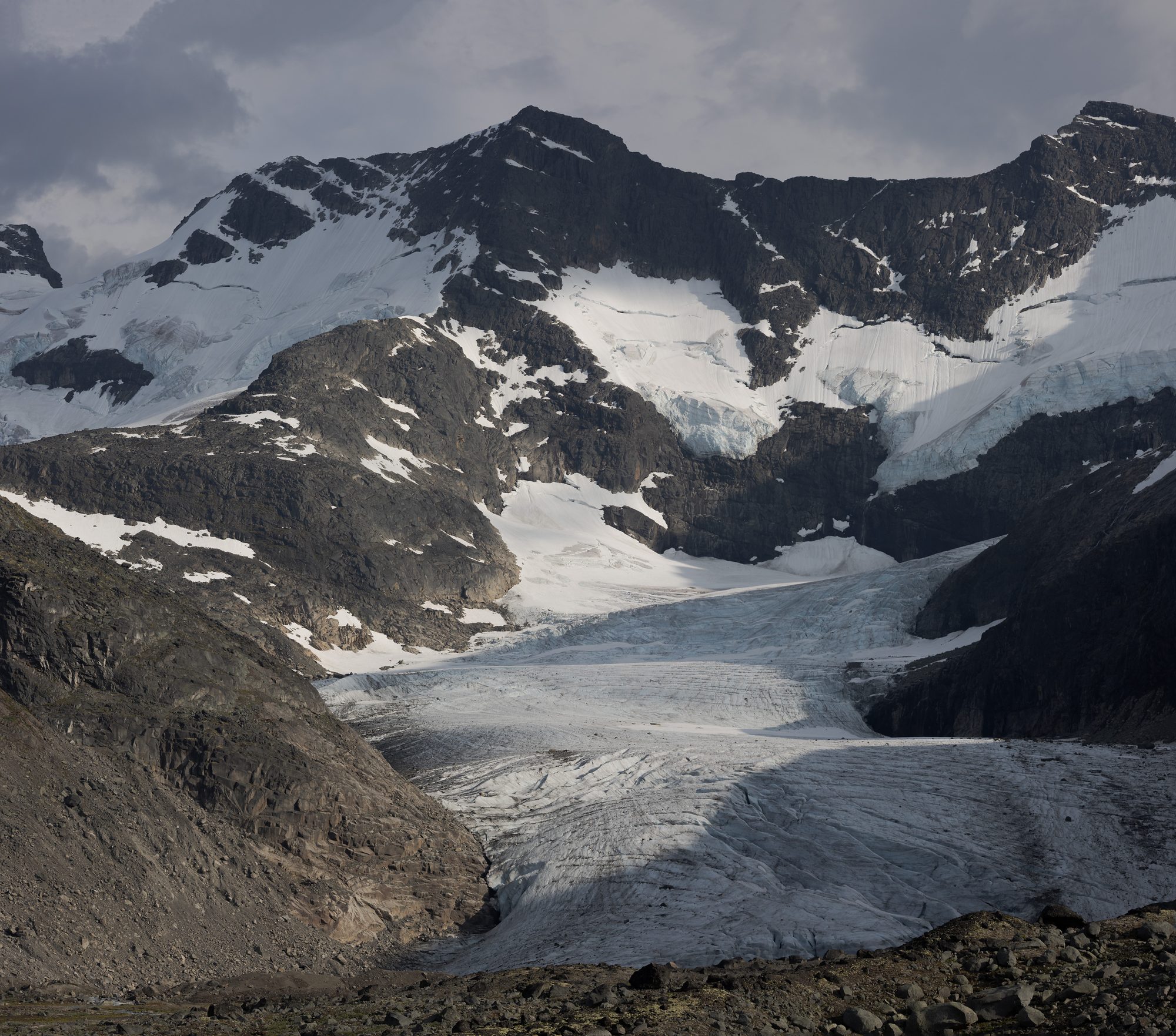 Styggedalsbreen spilling down between dark rock walls from a snow-covered ridge, grey moraine in the foreground