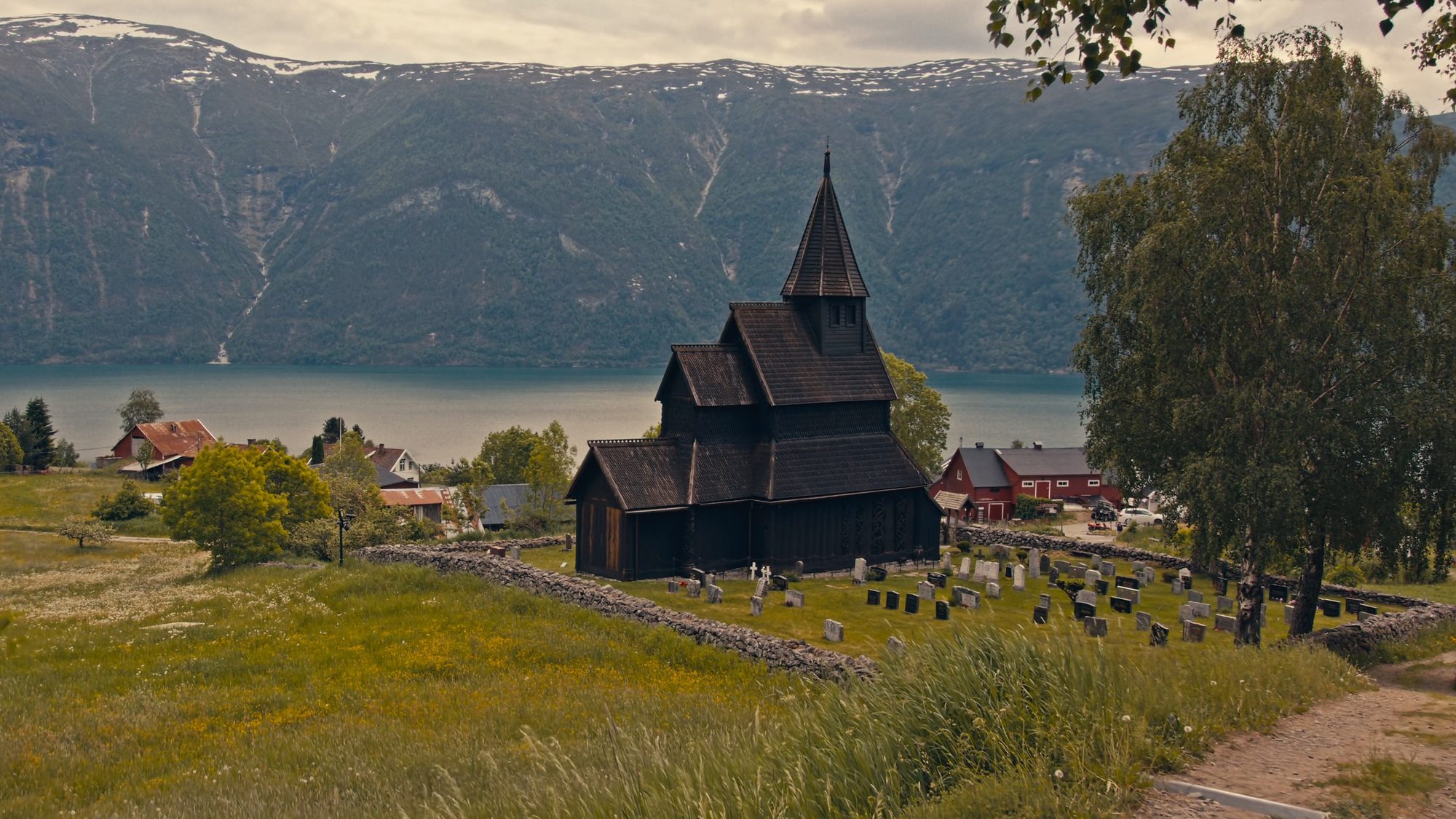 Urnes stave church on its slope above the Lustrafjord, the churchyard wall and gravestones around it and snow-streaked mountains across the water