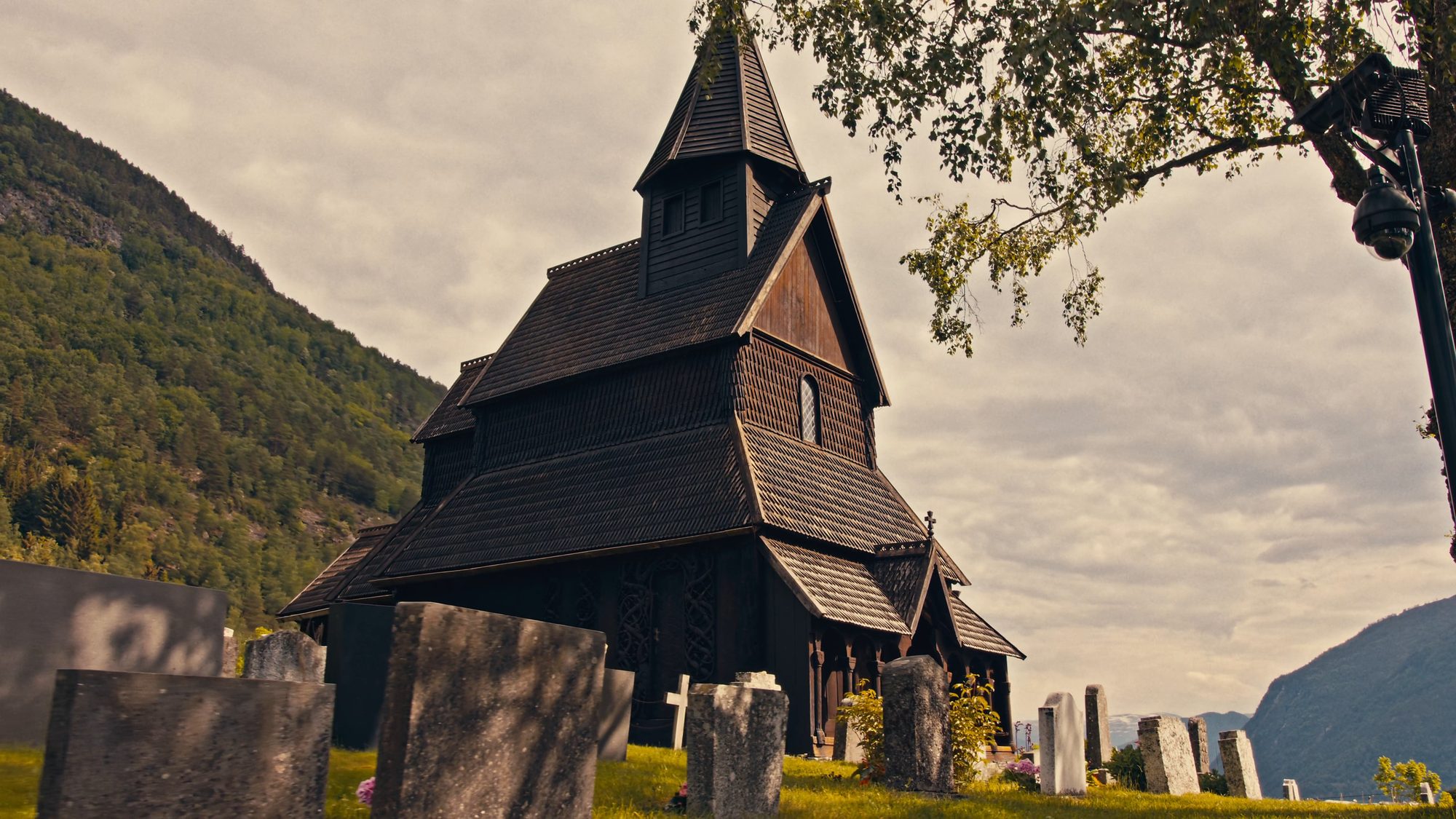 Urnes stave church seen from low among the gravestones, its dark tiered roofs and spire against a cloudy sky