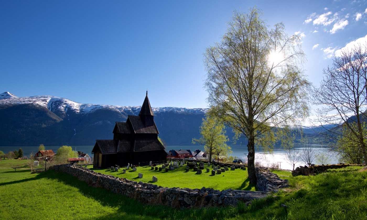 Urnes stave church inside its dry-stone churchyard wall on the green slope above the Lustrafjord, a birch in new leaf beside it with the sun behind, and snow-covered mountains across the water