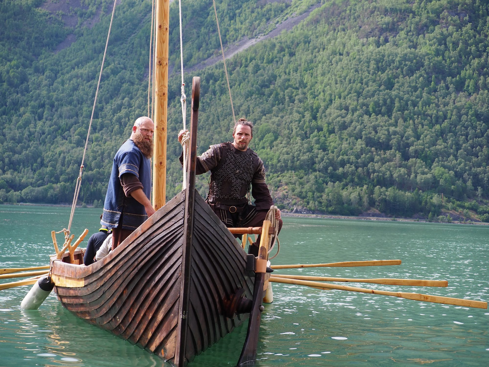 The Viking longship under oars on the fjord, two crew in period dress standing in the bow as she comes straight towards the camera, a wooded mountainside behind