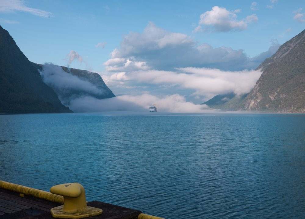 A yellow mooring bollard and the painted quay edge in the foreground, with the fjord running straight out between the mountain walls and a cruise ship standing off under a bank of low cloud