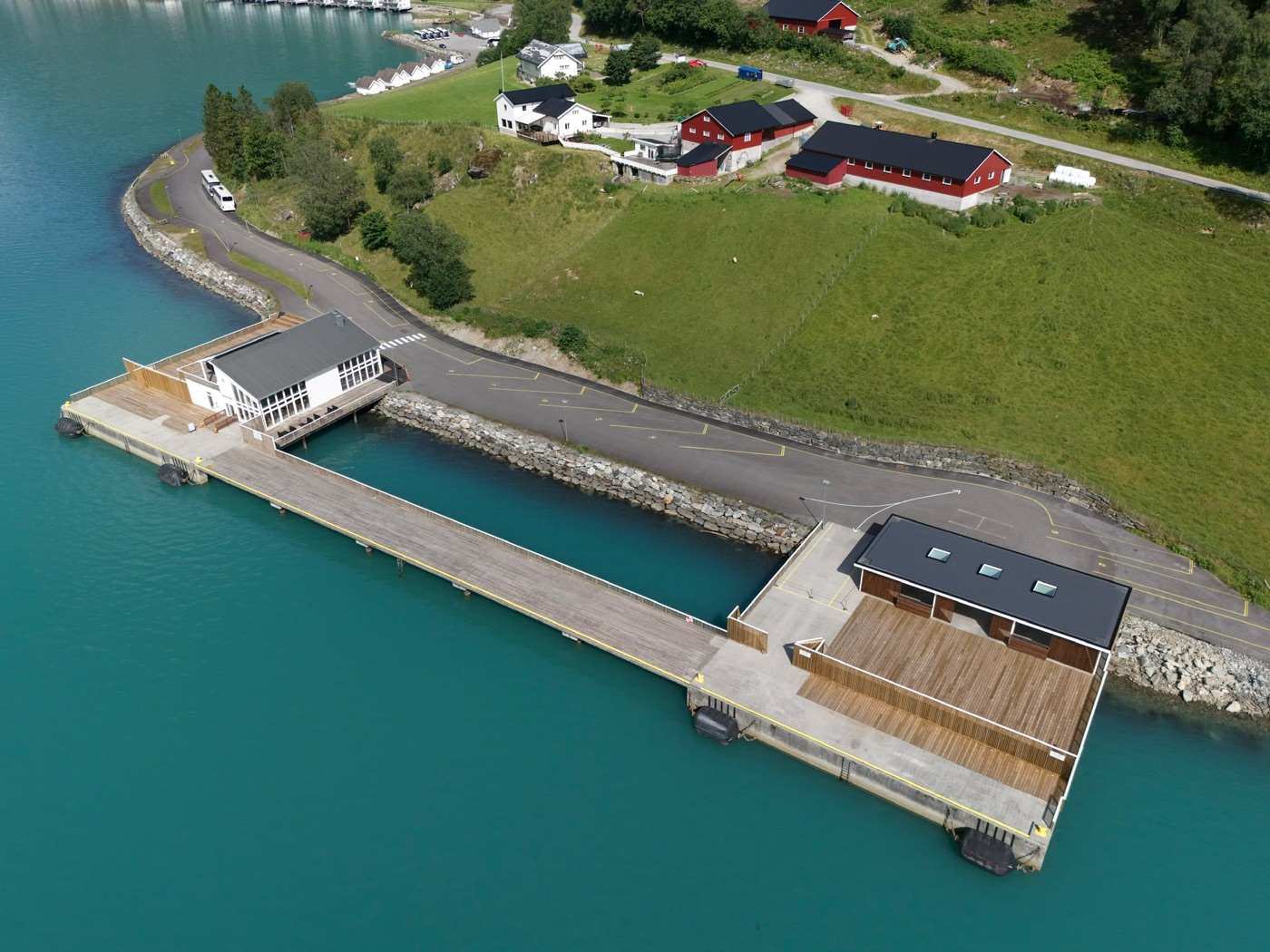 Oblique aerial view of the finished cruise pier: the concrete deck running out along the shore with black fenders down its face, the white gabled terminal building where it meets the land, a timber-clad building with a dark roof on the apron at the far end, and the marked road, green fields and red farm buildings behind