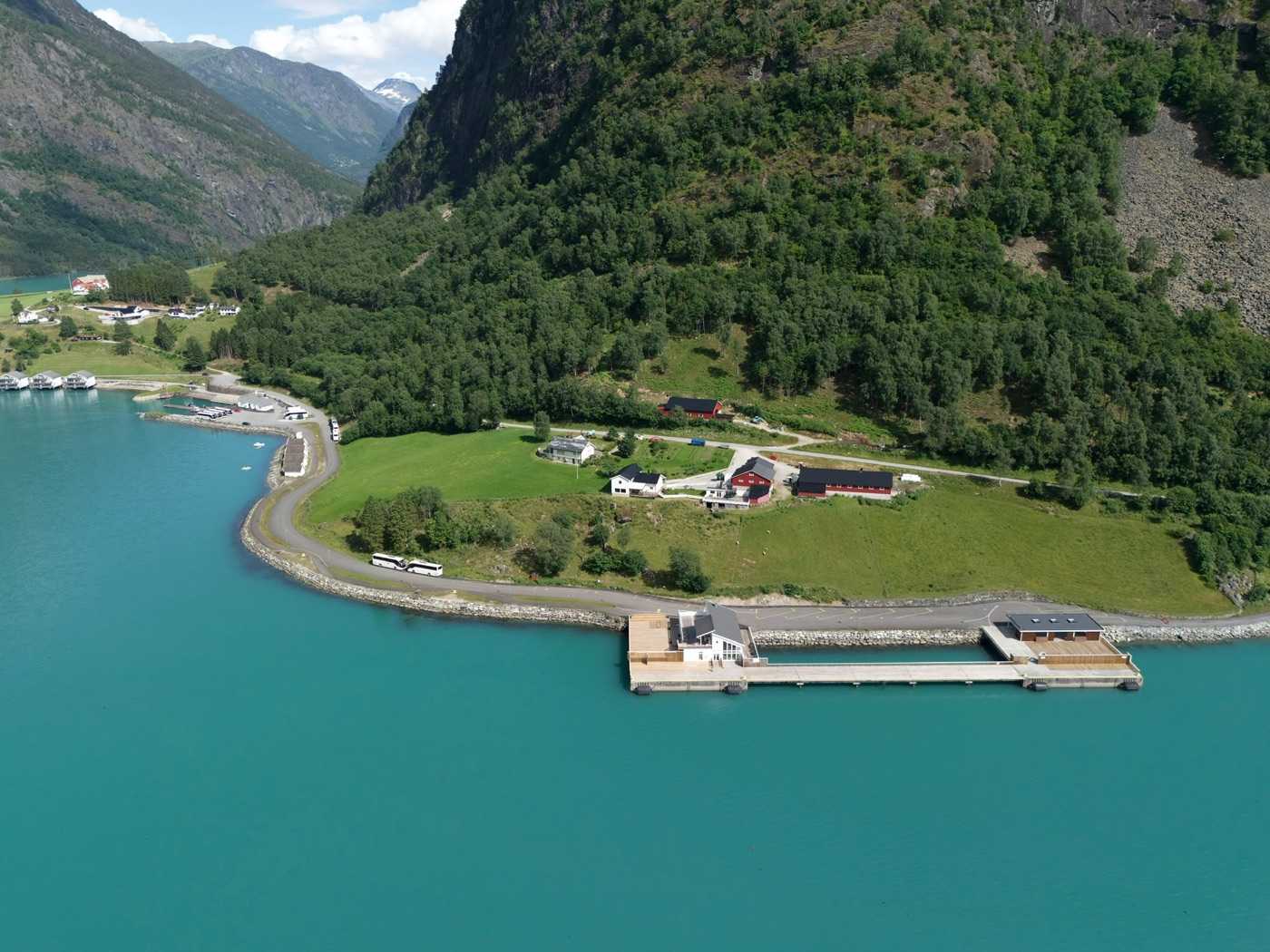 The cruise pier from the air in its setting: the deck and its two buildings on the point below a steep wooded mountainside, a coach standing on the shore road, the small-boat harbour further along the shore, and turquoise fjord water filling the foreground