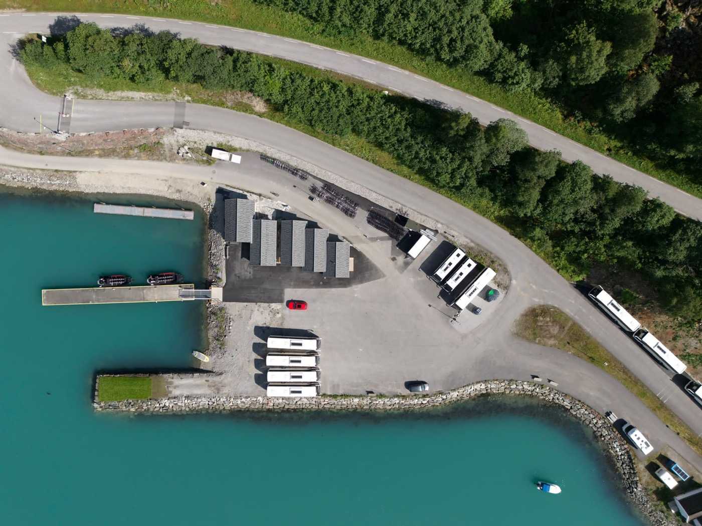 The tender pier from directly above: the floating pontoon with two RIBs alongside and its gangway to the shore, the paved yard behind it with coaches drawn up in rows, racks of bicycles along the road, and the barrier on the access road at the top of the frame
