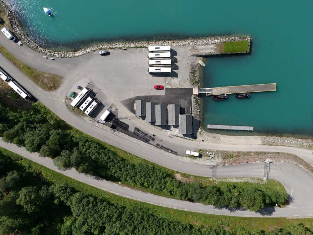 The coach park beside the tender pier seen from above, with coaches drawn up in rows on the gravel apron, more standing along the road beyond it, and the floating pontoon and its RIBs at the water's edge